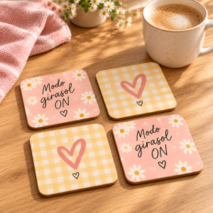 Cuatro posavasos: dos rosas con margaritas y la frase Modo girasol ON y dos de cuadros amarillos con corazón rosa, junto a una taza de café